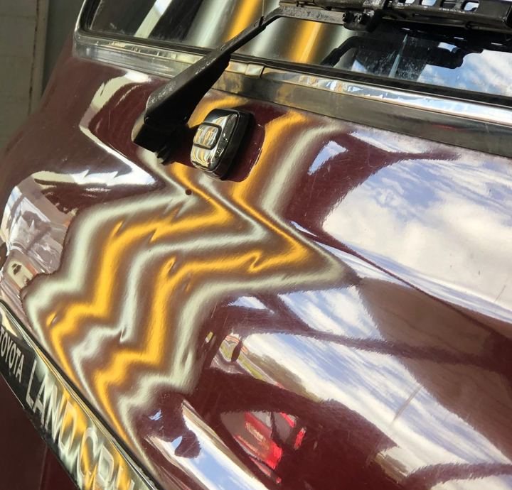 A burgundy Toyota LandCruiser panel under a striped light, the lines bending over a dent